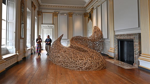 Two people looking at a large willow sculpture in a grand historic room with wooden floors and golden pillars